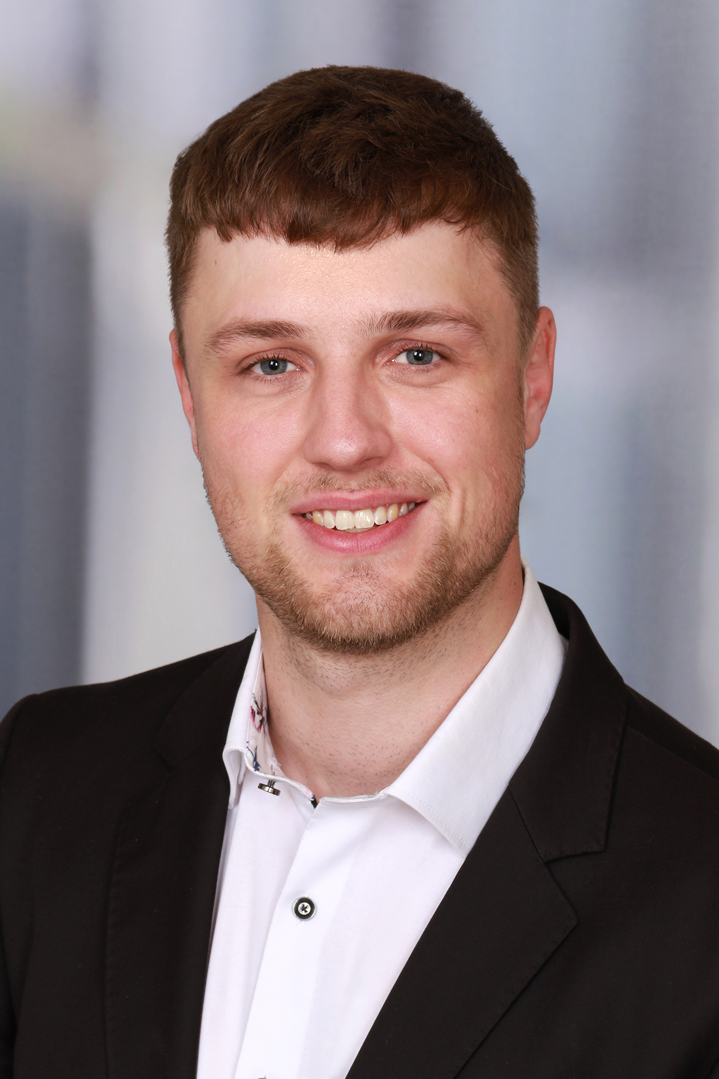 Professionelles Portrait von Marco Binder: lächelnd, dunkler Blazer, weißes Hemd, neutraler Studio-Hintergrund